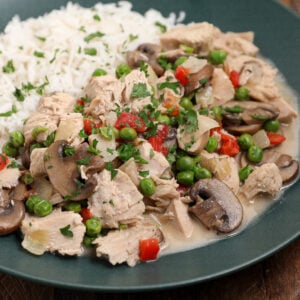 A plate of white rice served with a creamy Turkey À La King-style mixture of tuna chunks, mushrooms, peas, red peppers, onions, and garnished with chopped parsley.