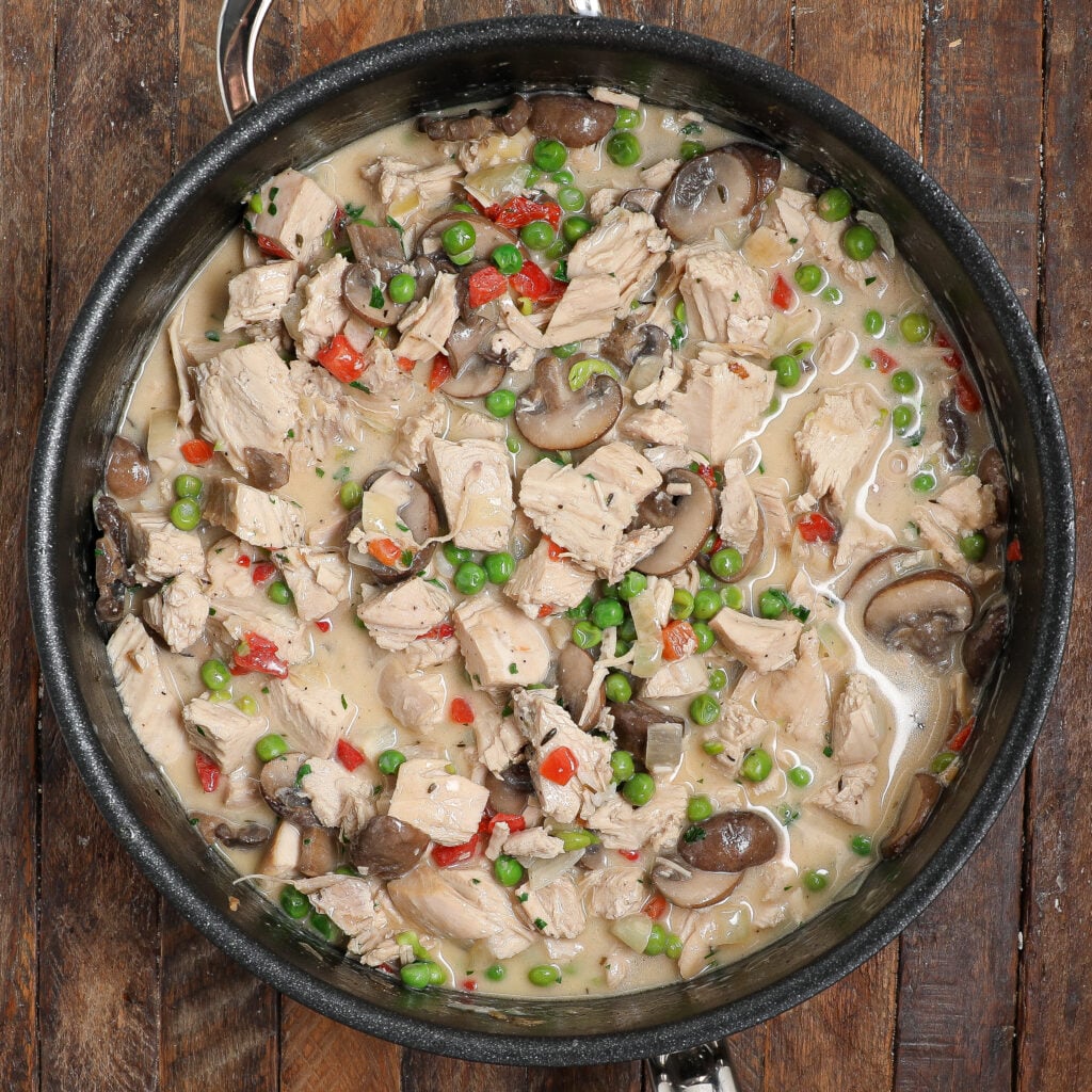 A skillet filled with a creamy Turkey À La King mixture of cooked chicken chunks, mushrooms, green peas, and red bell peppers, set on a rustic wooden surface.