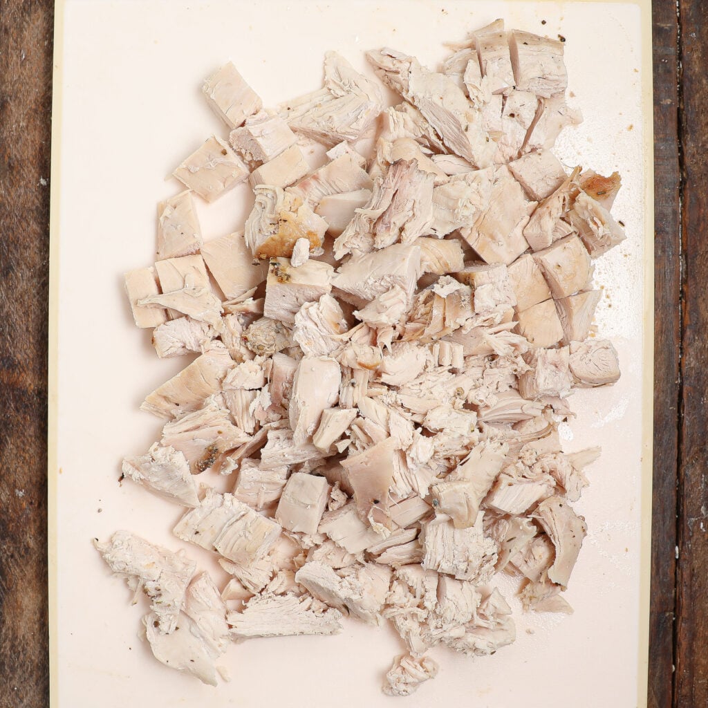 Chopped cooked chicken breast pieces, perfect for dishes like Turkey À La King, are spread out on a white cutting board, placed on a rustic wooden surface.