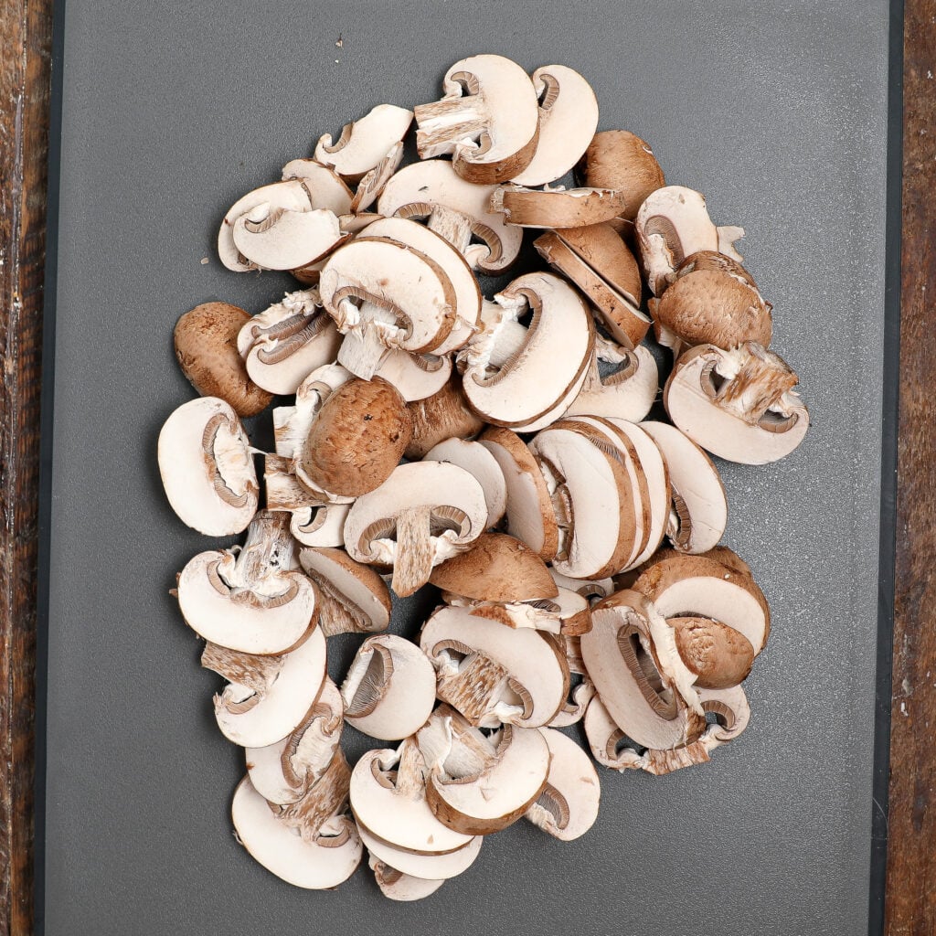 A pile of sliced brown mushrooms, perfect for Turkey À La King, is spread out on a dark gray cutting board with a wooden edge visible on both sides.