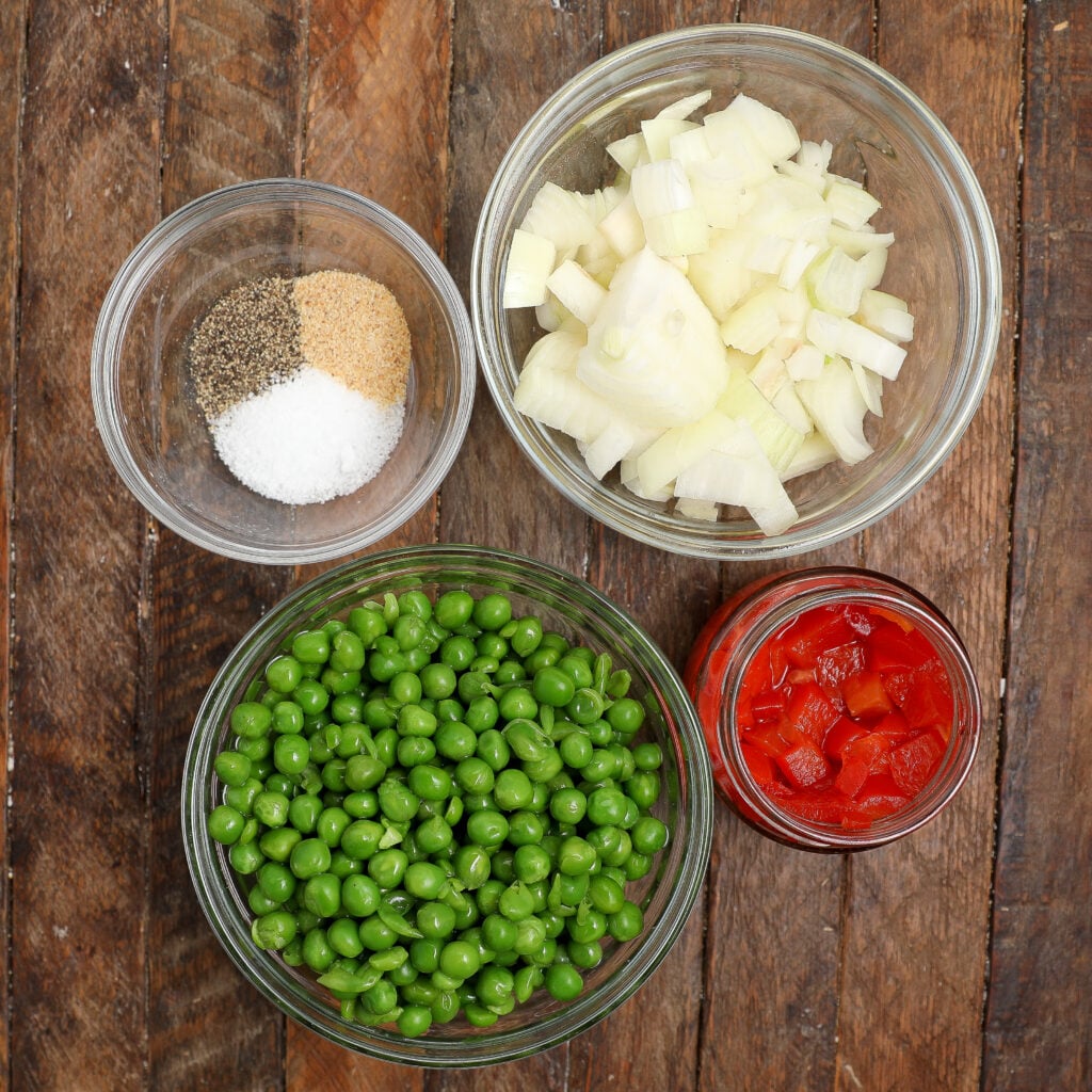 Four glass bowls on a wooden surface display key Turkey À La King ingredients: spices and salt, chopped onions, diced tomatoes, and green peas.