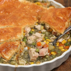 A close-up of a Turkey Pot Pie with a golden, flaky crust. The pie is partially cut open, revealing chunks of turkey, peas, carrots, corn, and celery in a creamy filling. A serving spoon is inside the dish.