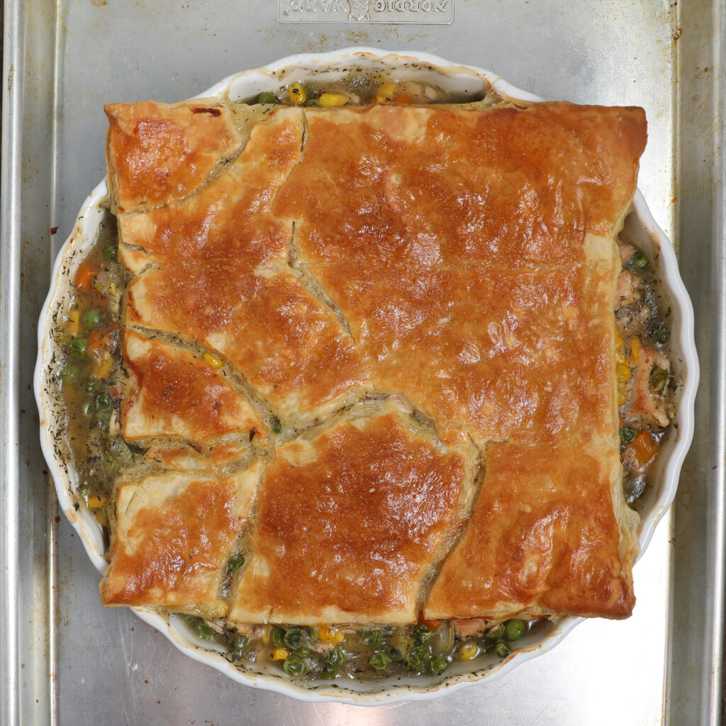 A golden-brown, square-topped Turkey Pot Pie with flaky crust sits in a round white dish, slightly overflowing with visible vegetables and sauce, placed on a silver baking tray.