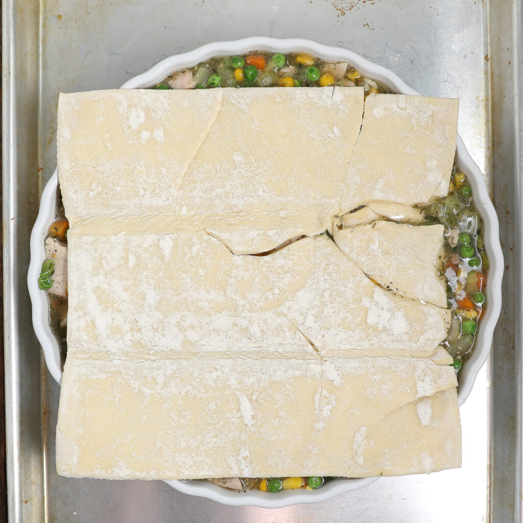 Unbaked Turkey Pot Pie in a white dish, topped with uneven pieces of raw pastry dough, shows its hearty vegetable filling with peas, carrots, and corn underneath, all resting on a metal baking sheet.