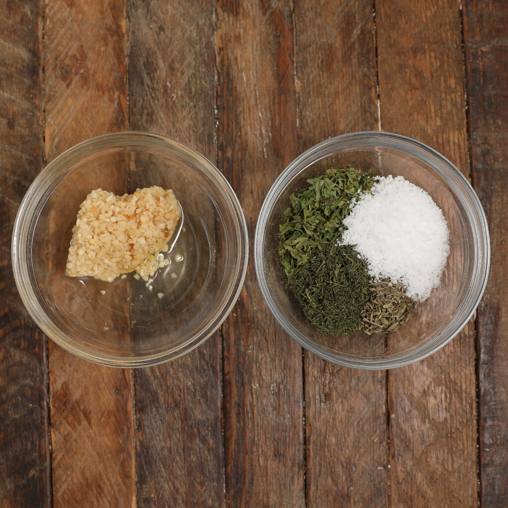 Two glass bowls on a wooden surface: one contains minced garlic, the other holds dried herbs and a mound of coarse salt, arranged in sections—essential aromatics for preparing a flavorful Turkey Pot Pie.