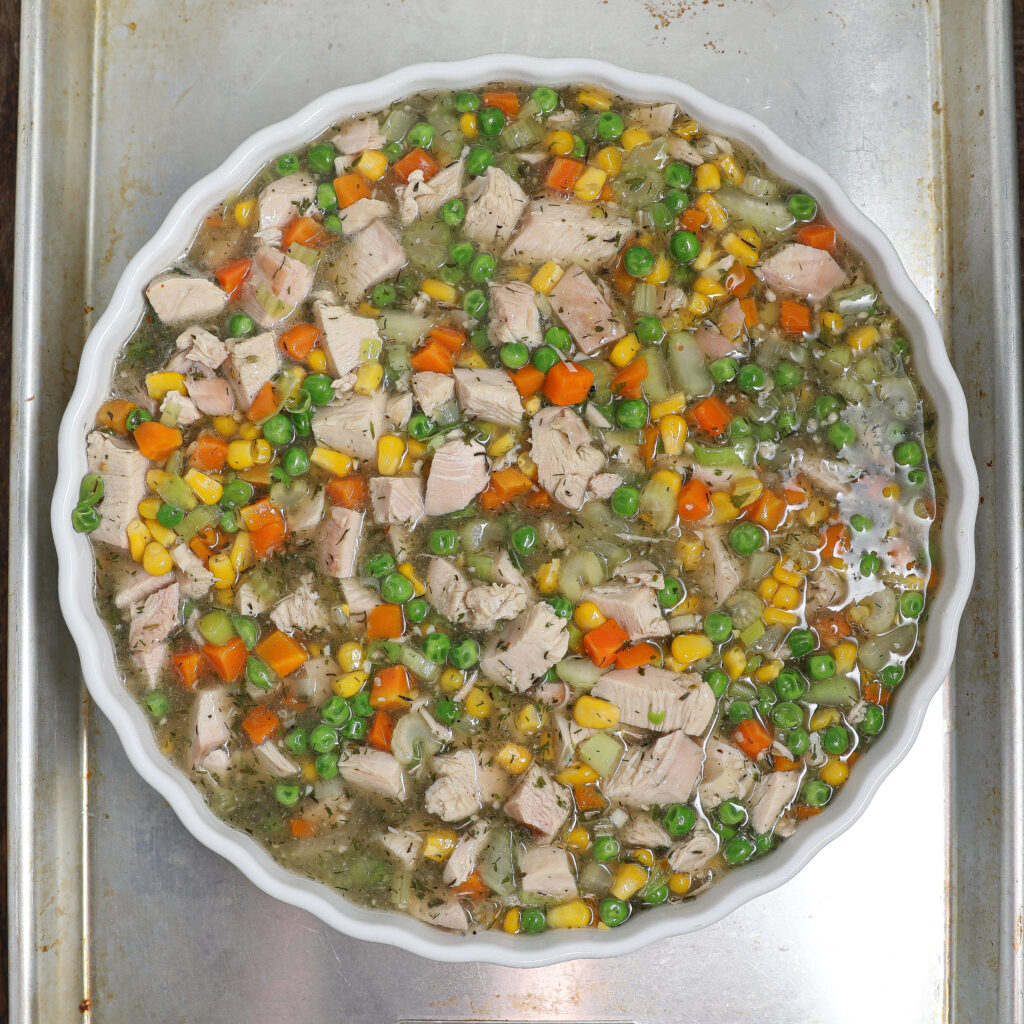 A round white dish filled with a mixture of cubed chicken, peas, corn, carrots, potatoes, and broth sits on a metal baking sheet—perfect for creating a comforting Turkey Pot Pie or classic chicken pot pie.