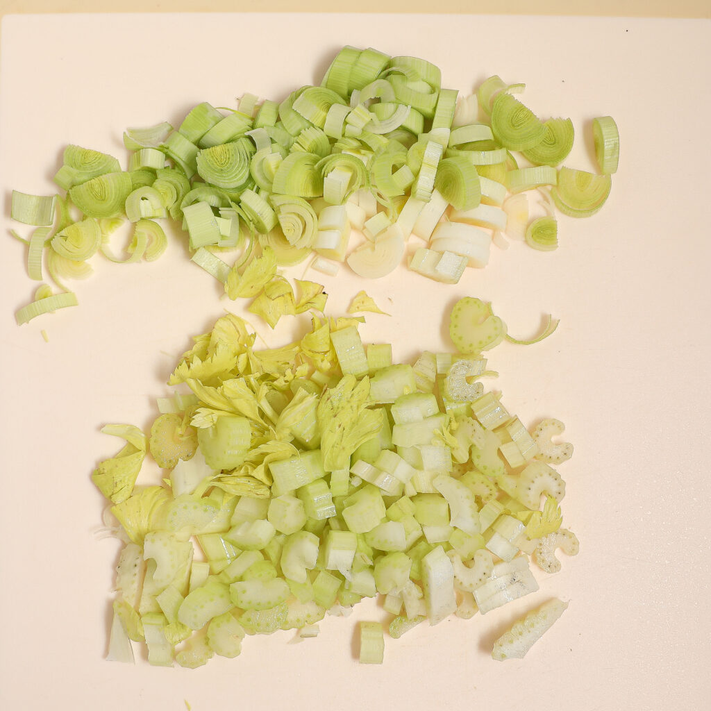 Chopped leeks and celery, perfect for a Turkey Pot Pie, are arranged separately on a white cutting board. The leeks are light green and white, while the celery pieces are pale green with visible leaves.