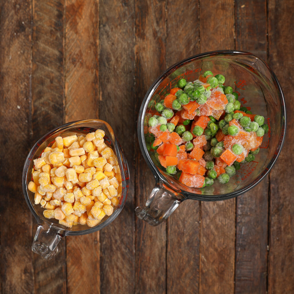 Two glass measuring cups on a wooden surface, one filled with frozen corn and the other with a mix of frozen green peas and diced carrots—perfect veggie prep for making a classic Turkey Pot Pie.