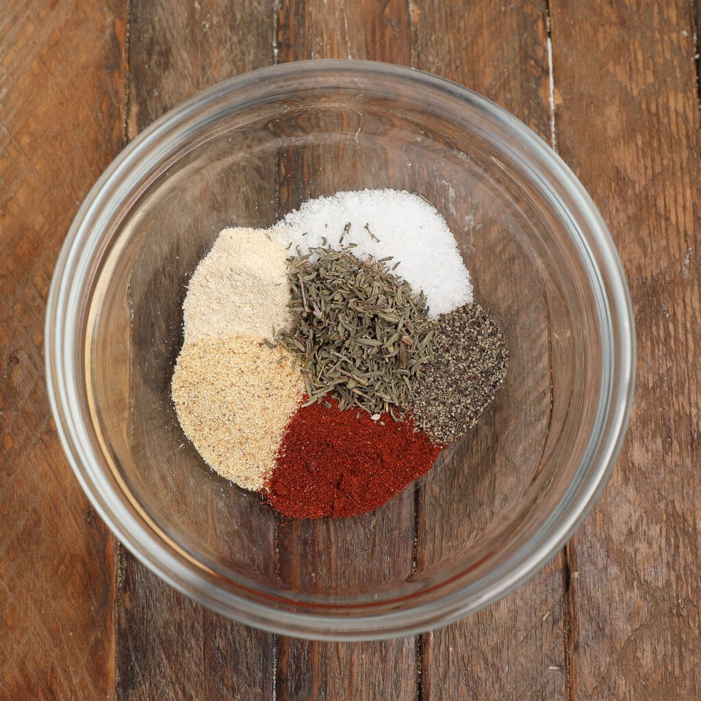 A glass bowl on a wooden surface contains small piles of various spices, including paprika, black pepper, dried herbs, onion powder, garlic powder, and salt—perfect for seasoning a hearty turkey stew.