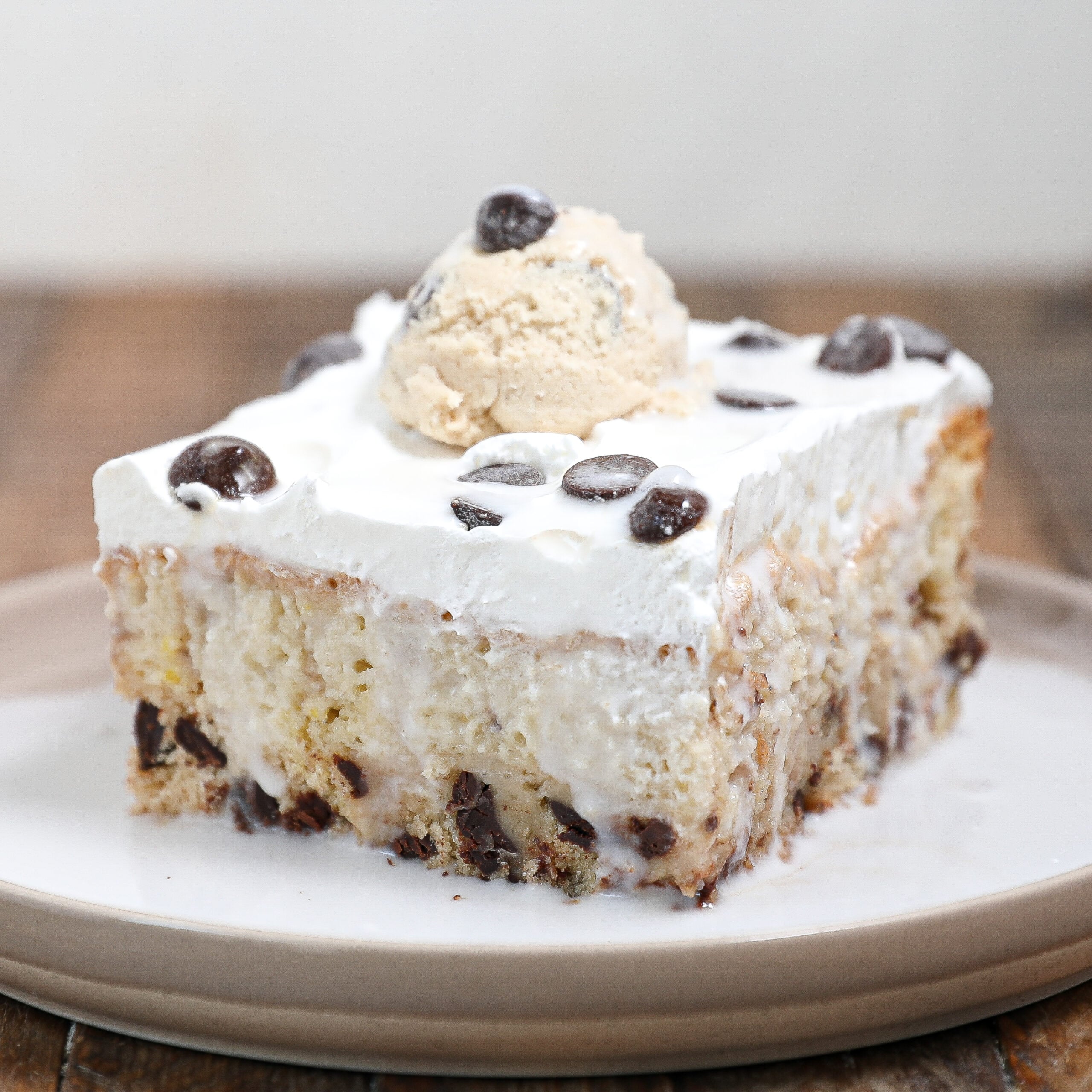 A square slice of Chocolate Chip Cookie Dough Tres Leches cake with chocolate chips, topped with whipped cream, a scoop of cookie dough, and more chocolate chips, served on a beige plate.