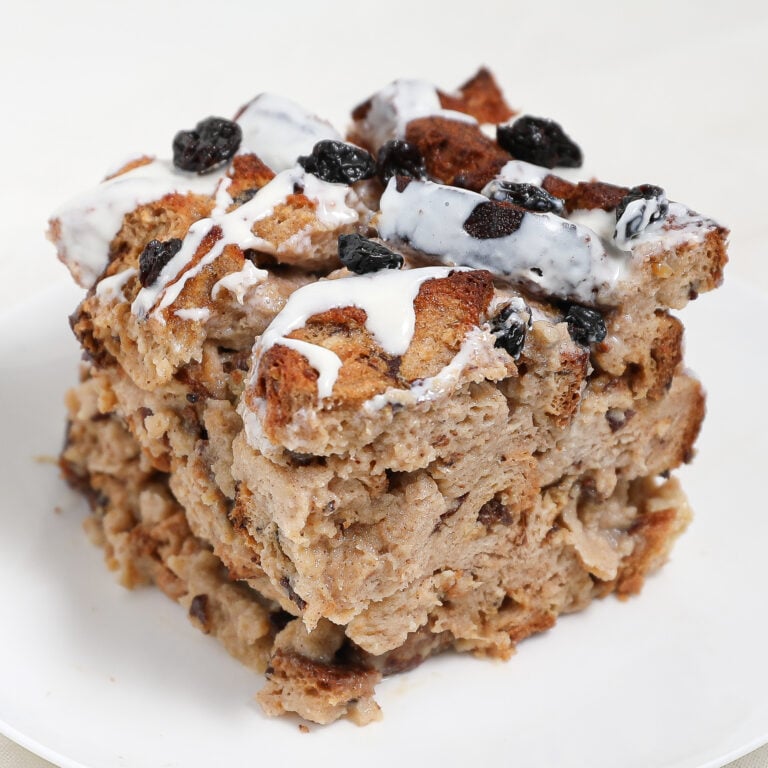 A thick square slice of Cinnamon Raisin Bread Pudding topped with raisins and drizzled with white icing sits on a white plate. The bread is golden brown and appears moist and dense.