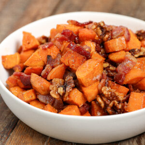 A white bowl filled with Maple Roasted Sweet Potato with Bacon & Walnuts sits on a wooden surface, featuring crispy bacon pieces and glazed walnuts.