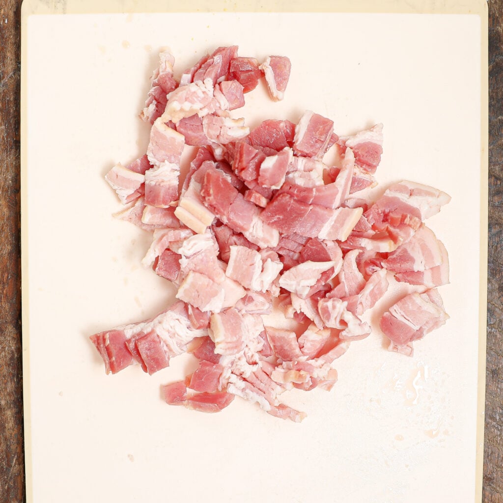Small cubes of raw bacon are scattered on a white cutting board, ready to be added to a Maple Roasted Sweet Potato with Bacon & Walnuts dish.