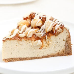 A slice of White Chocolate Macadamia Nut Cheesecake with caramel sauce, peanuts, and a drizzle of white icing, served on a white plate.
