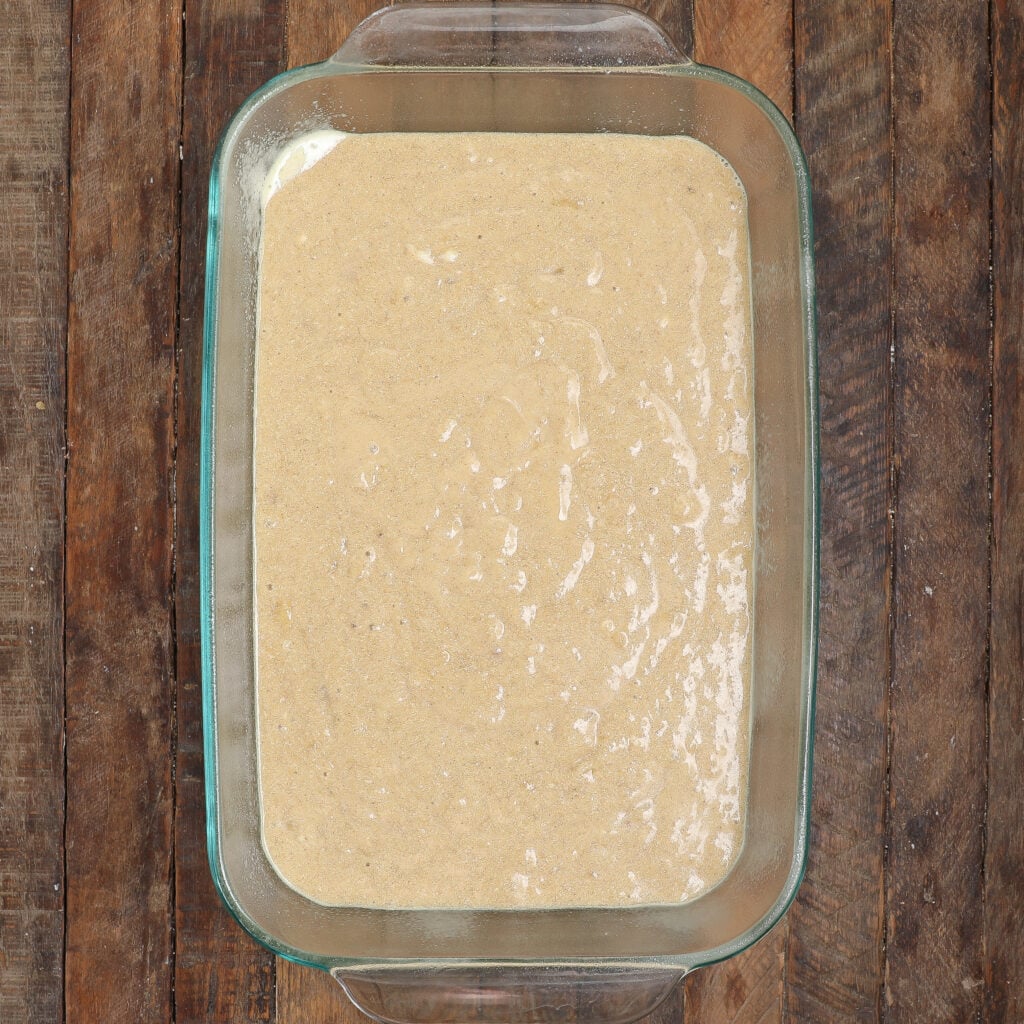 A glass baking dish filled with an even layer of sticky banana bread pudding batter sits on a rustic wooden surface.