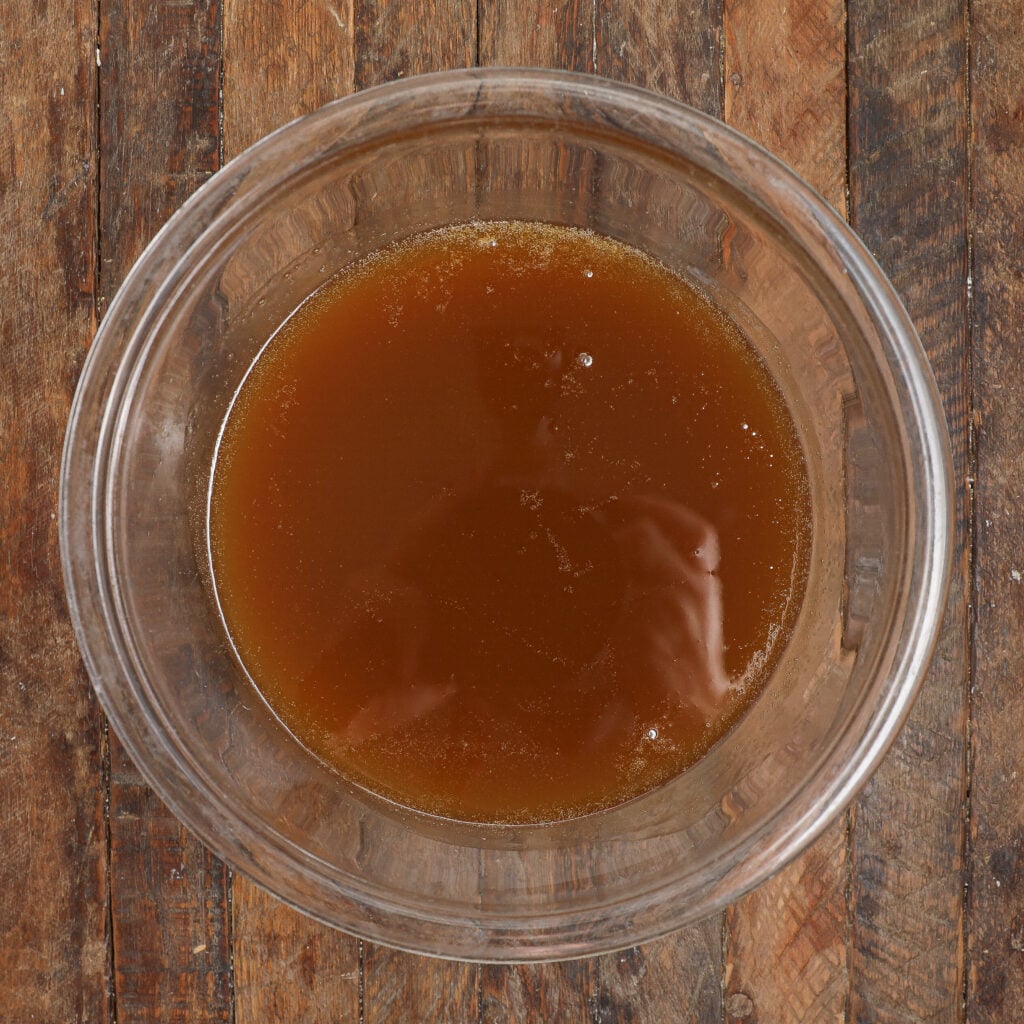 A glass bowl filled with brown liquid, possibly broth or stock, sits on a rustic wooden surface—perfect for pairing with a serving of sticky banana bread pudding.
