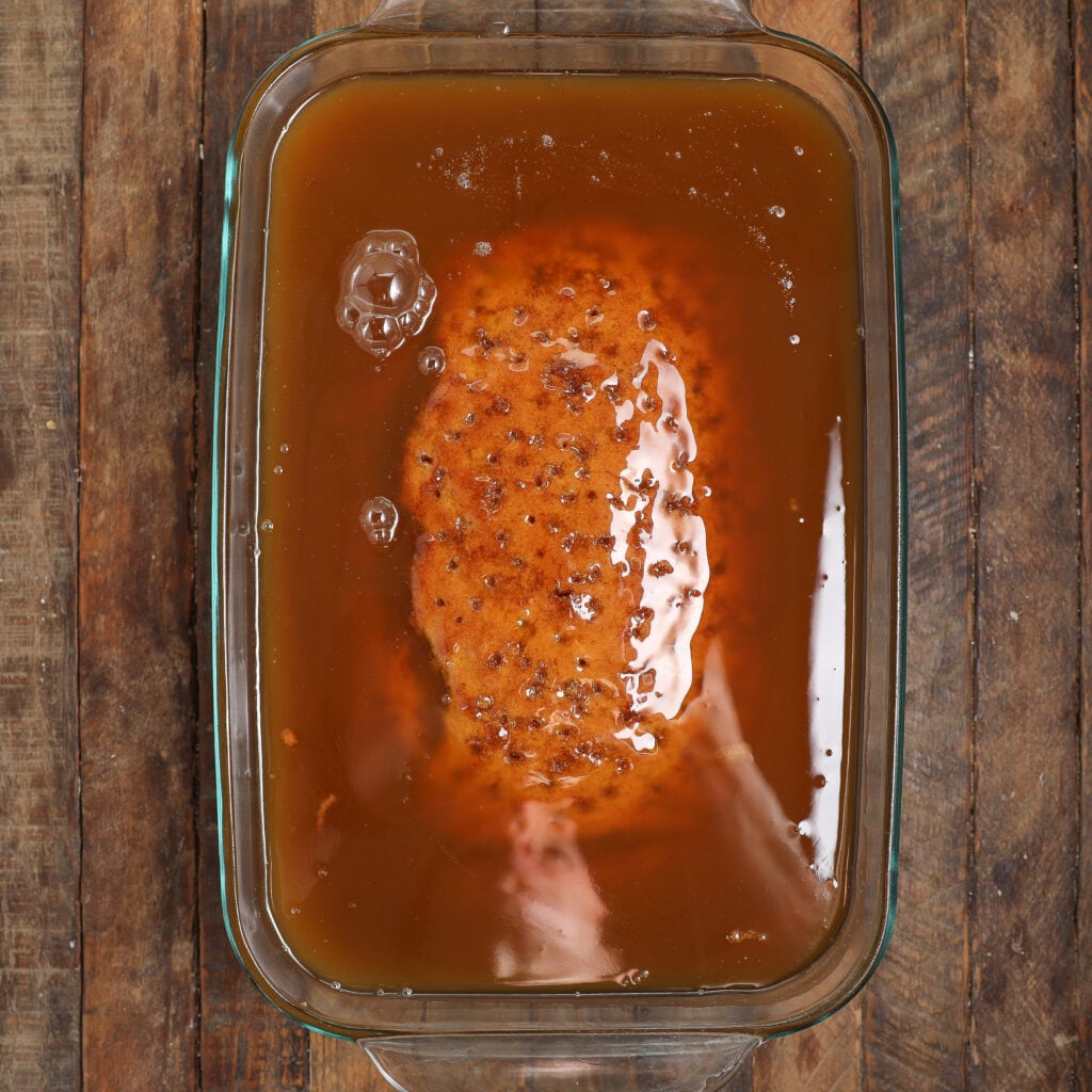 A raw roast is soaking in a brown liquid brine inside a glass baking dish on a wooden surface, reminiscent of the rich flavors found in sticky banana bread pudding.