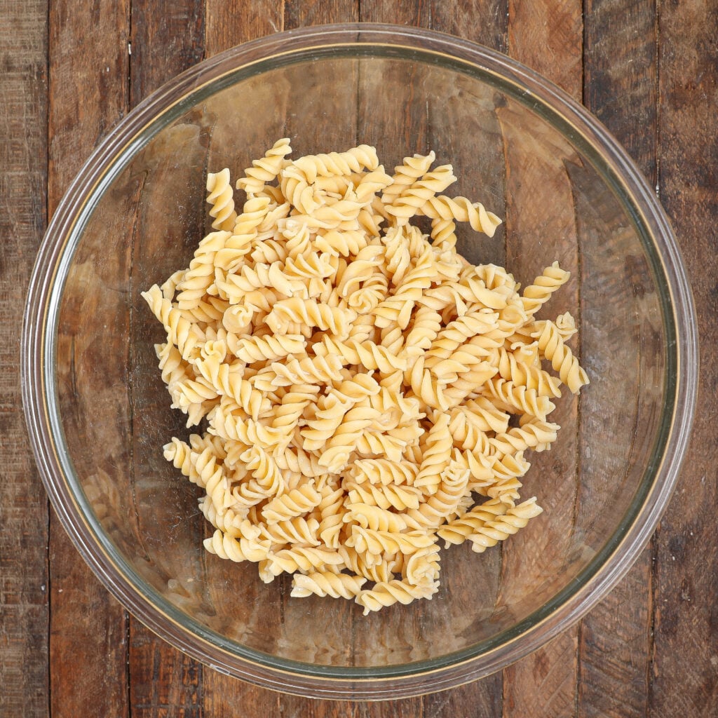 A clear glass bowl filled with uncooked rotini pasta sits on a rustic wooden surface, ready to be transformed into a delicious Taco Pasta Salad.
