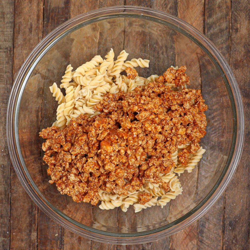A glass bowl filled with cooked rotini pasta and seasoned ground meat sauce, creating a hearty Taco Pasta Salad, placed on a rustic wooden surface.