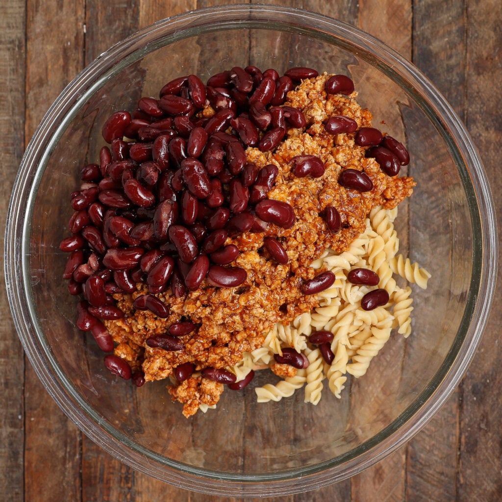 A glass bowl on a wooden surface contains dry rotini pasta, kidney beans, and a crumbled meat mixture—an inviting start for Taco Pasta Salad, viewed from above.