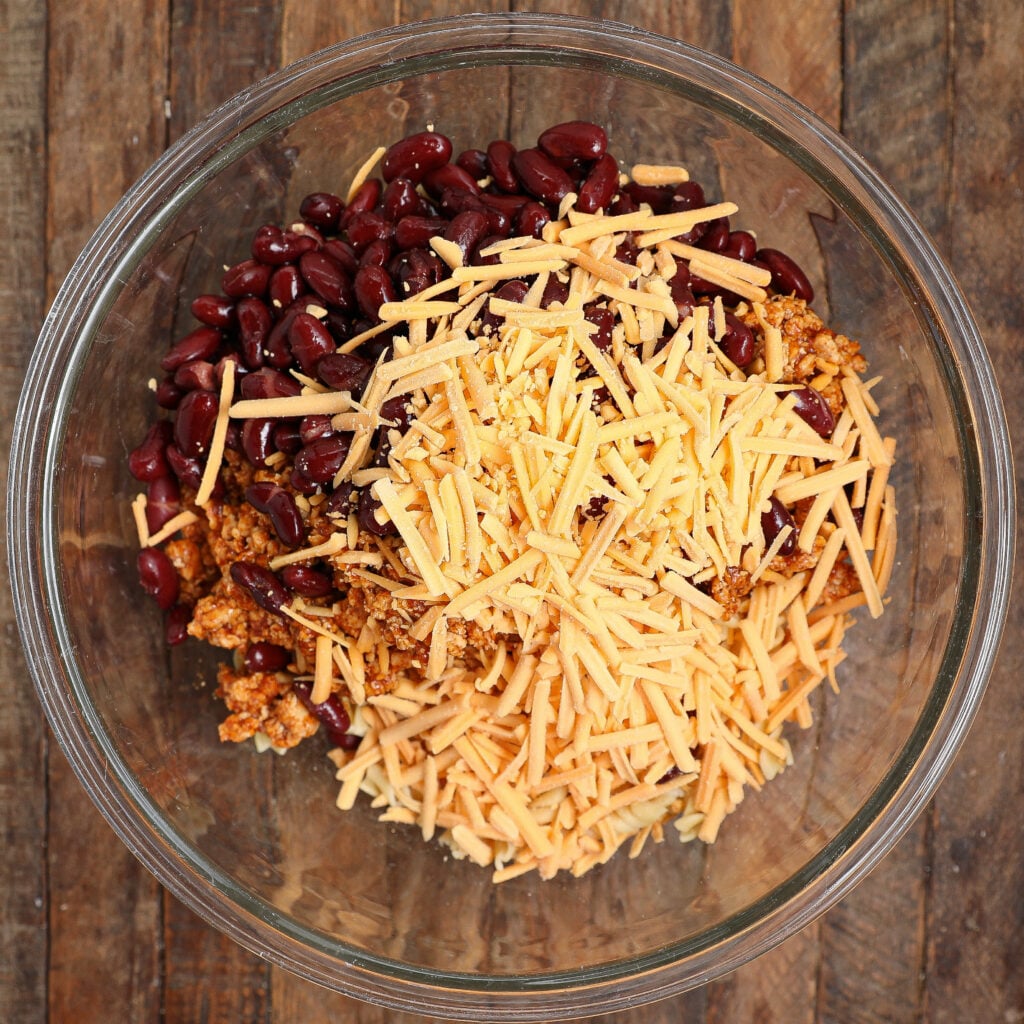 A glass bowl on a wooden surface contains kidney beans, shredded cheddar cheese, and seasoned ground meat, ready to be mixed for a flavorful Taco Pasta Salad.