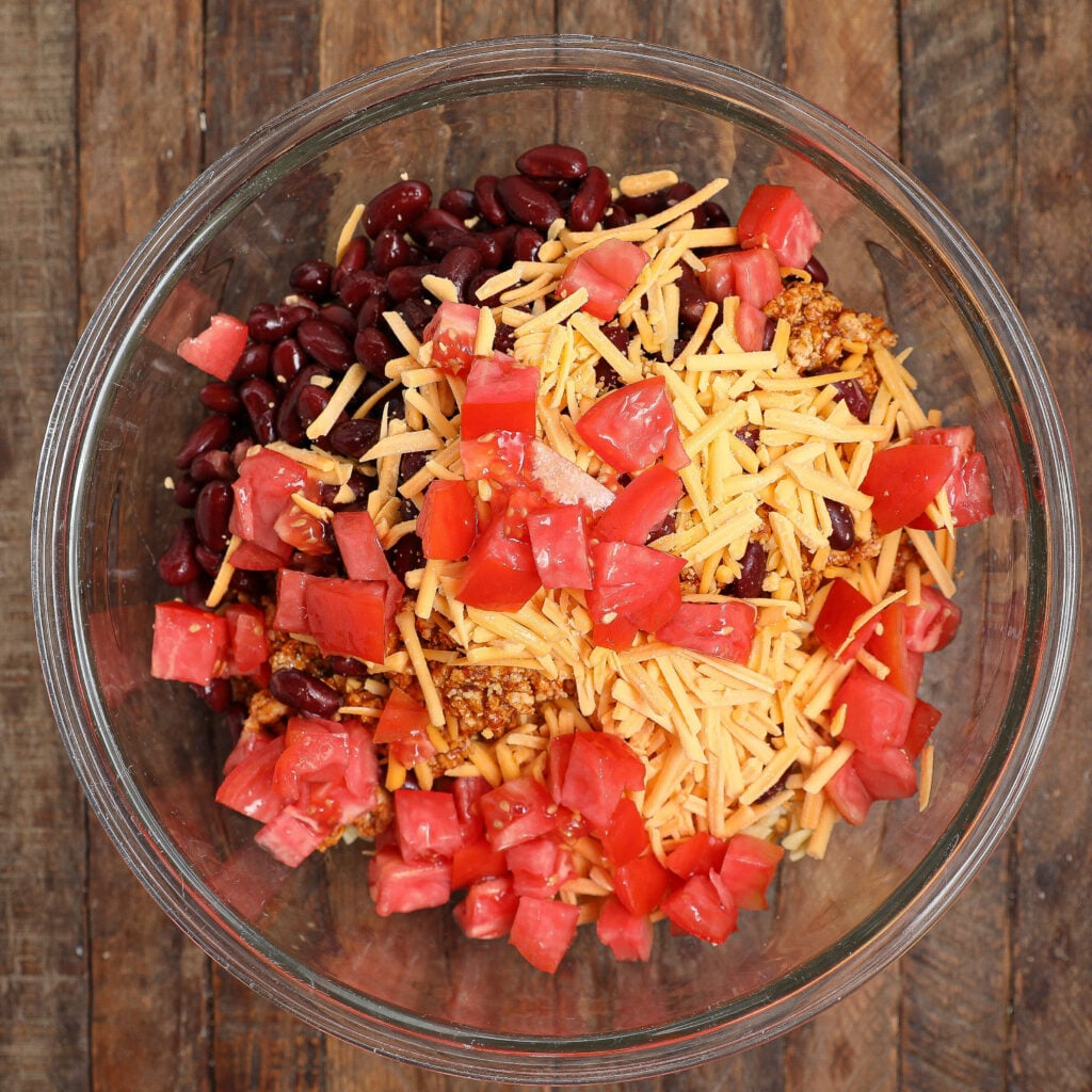 A clear glass bowl on a wooden surface filled with kidney beans, shredded cheddar cheese, diced tomatoes, and taco-seasoned ground meat creates a flavorful Taco Pasta Salad perfect for any gathering.