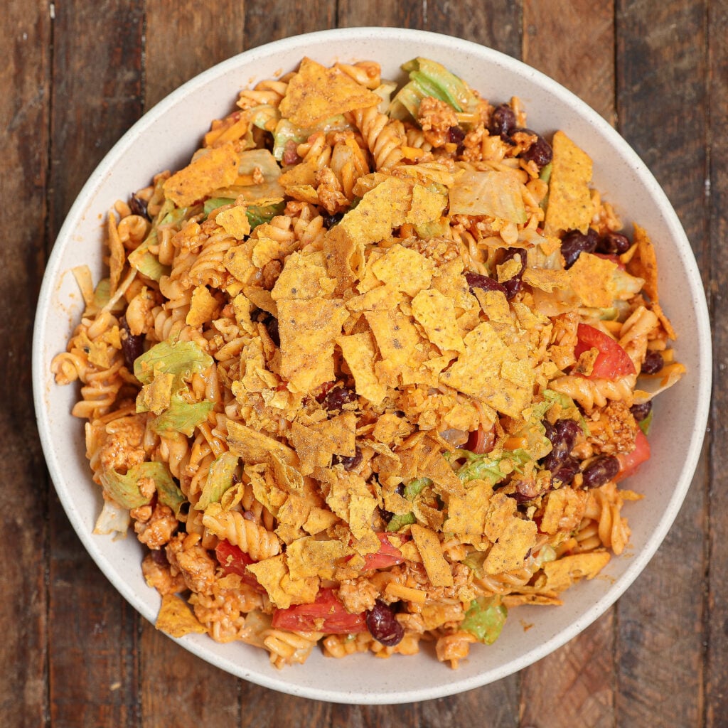 A bowl of Taco Pasta Salad with rotini pasta, ground meat, kidney beans, lettuce, tomatoes, and crushed tortilla chips on top, served on a wooden table.