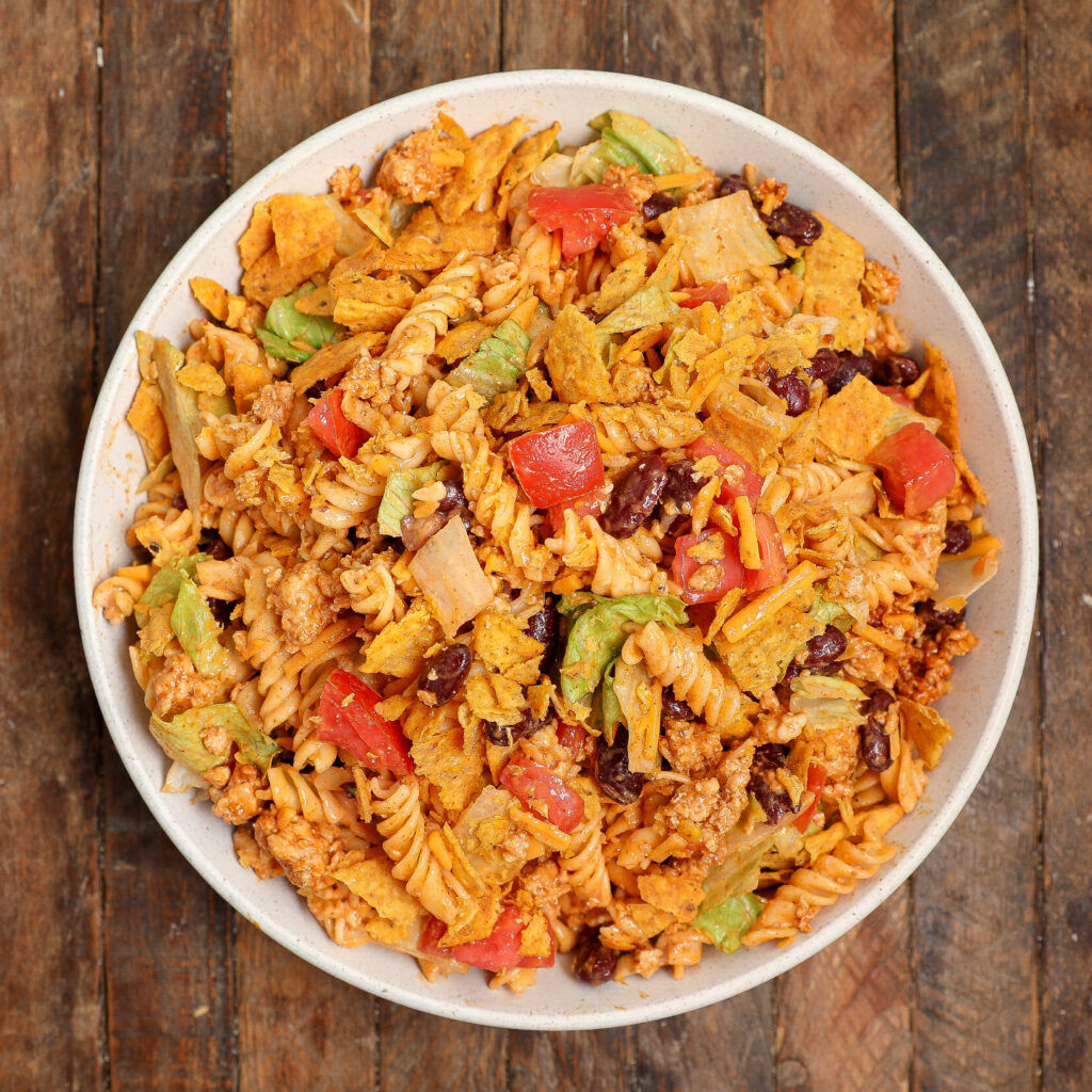 A bowl of Taco Pasta Salad with rotini pasta, ground meat, chopped lettuce, tomatoes, kidney beans, cheese, and crumbled tortilla chips is served on a rustic wooden surface.