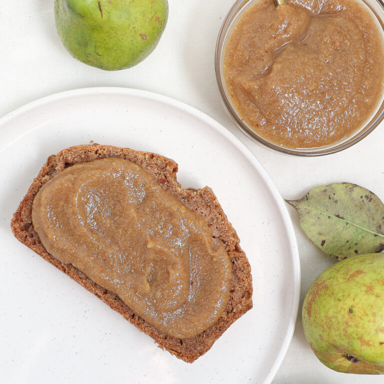 A slice of bread spread with pear butter sits on a white plate, next to a glass bowl filled with more pear butter. Whole pears and leaves are arranged nearby on a white cloth, highlighting the homemade charm of this pear butter treat.