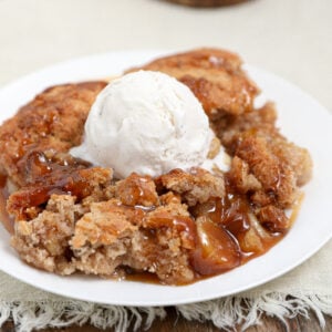 A plate of warm banana caramel cobbler topped with a scoop of vanilla ice cream, featuring golden-brown crumbly crust, gooey caramelized bananas and apples, and melted ice cream on a white plate.