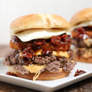 A close-up of a gourmet breakfast smashburger with two beef patties, melted cheese, crispy hash brown, fried egg, and caramelized onions or bacon on a toasted bun, served on a white plate. Another burger is blurred in the background.