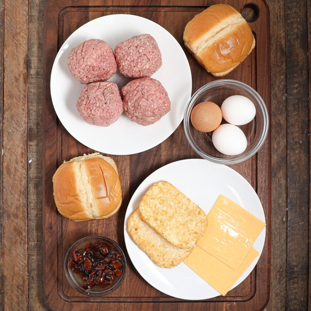 Top-down view of a wooden board with plates holding ground beef patties, hash browns, American cheese slices, burger buns, eggs in a bowl, and a small dish of caramelized onions—perfect ingredients for breakfast smashburgers.