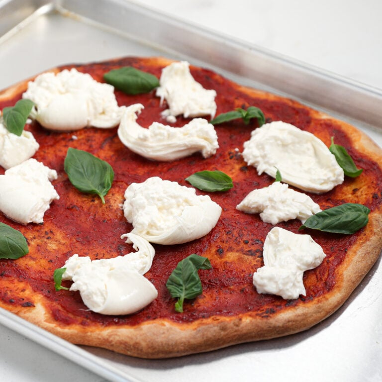 A rectangular pizza crust topped with tomato sauce, creamy burrata cheese, and scattered basil leaves rests on a metal baking tray.