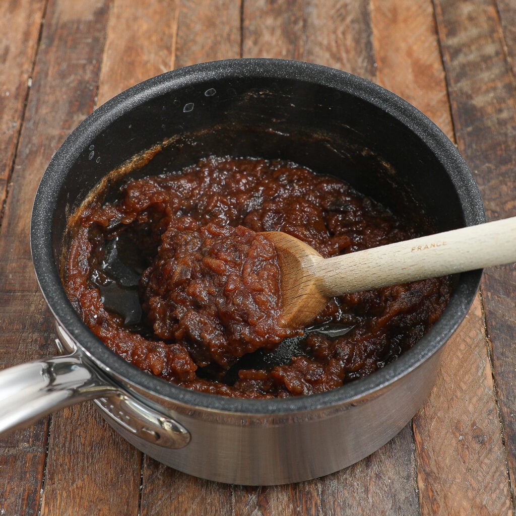 A saucepan filled with thick, dark brown apple butter—perfect for Caramel Apple Sticky Pudding Cake—is being stirred with a wooden spoon, resting on a rustic wooden surface.