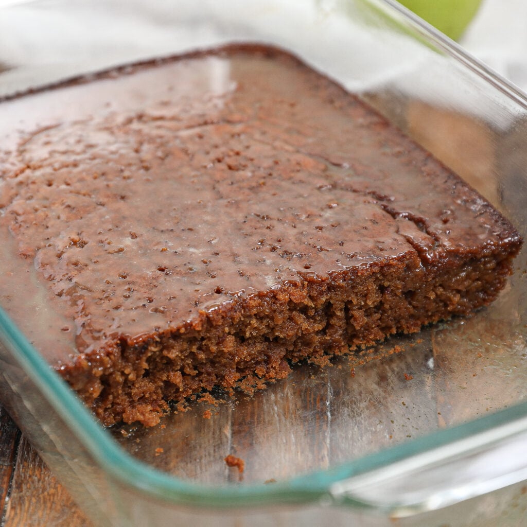 A glass baking dish with a partially cut, moist Caramel Apple Sticky Pudding Cake topped with a shiny glaze, showing its soft texture and crumb.