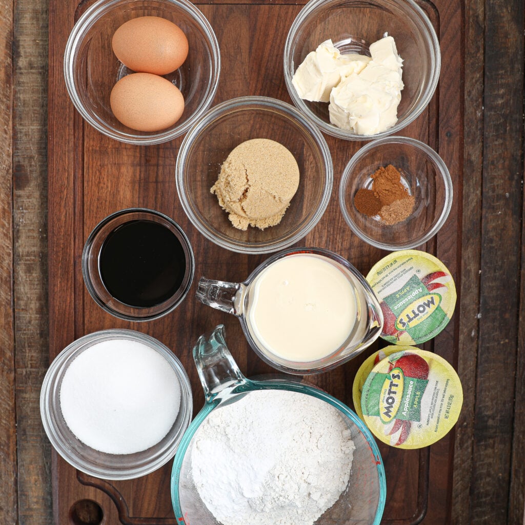 A wooden board holds bowls of eggs, butter, sugars, flour, vanilla, spices, sweetened condensed milk, and applesauce—ingredients lined up to make a delicious Caramel Apple Sticky Pudding Cake.