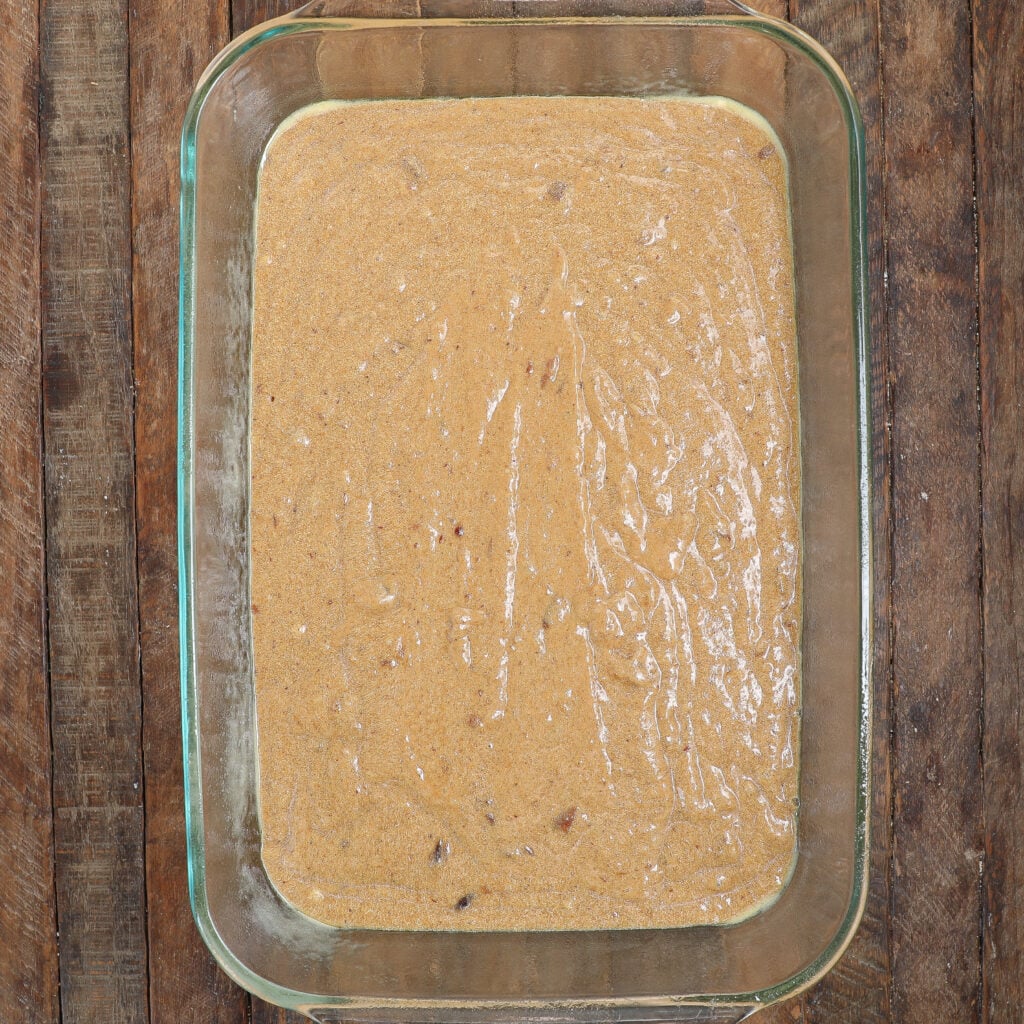 A glass baking dish filled with uncooked Caramel Apple Sticky Pudding Cake batter rests on a rustic wooden surface. The smooth, light brown batter contains small visible chunks mixed in.