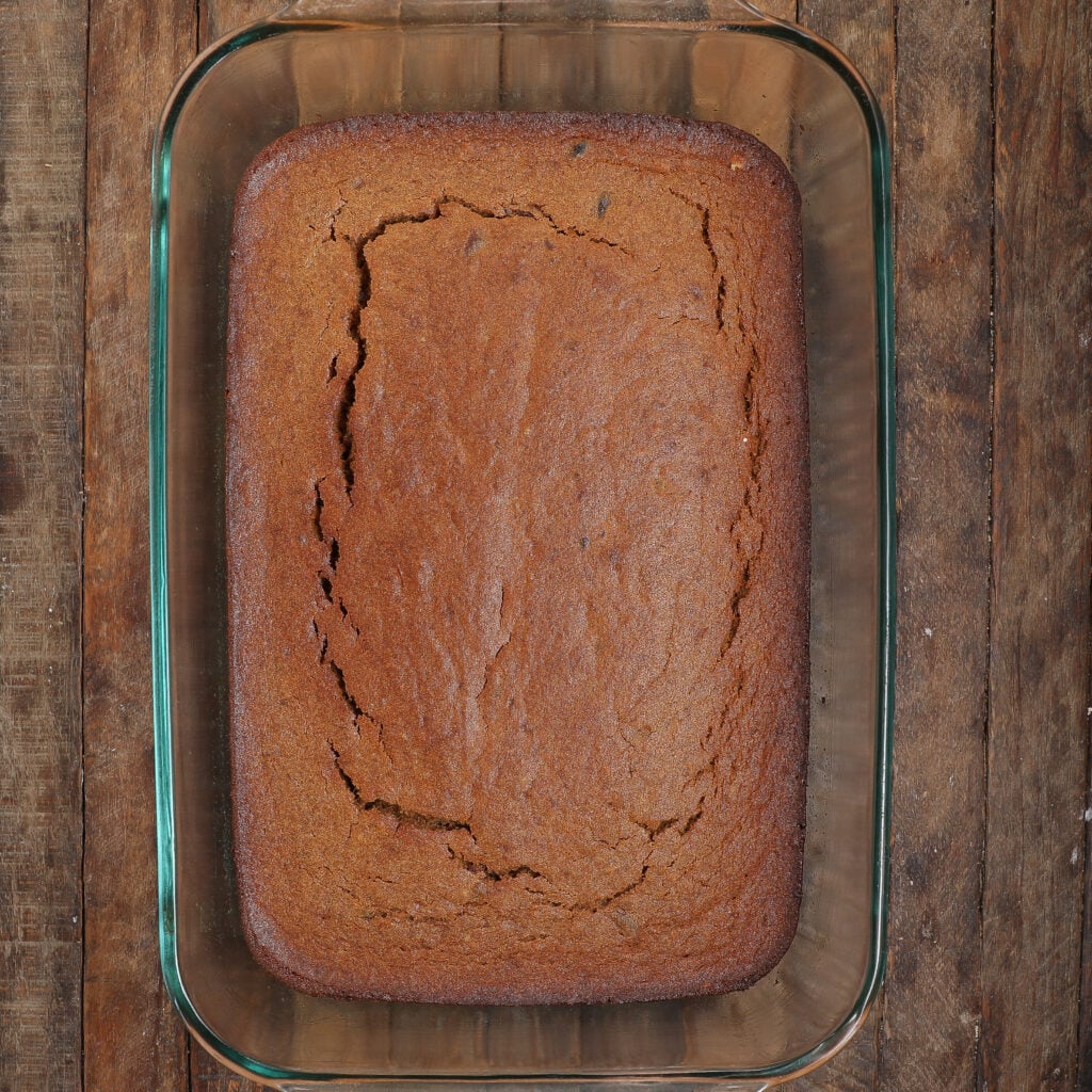 A rectangular, golden-brown loaf of baked cornbread sits in a clear glass baking dish on a rustic wooden table, its cracked top reminiscent of a freshly baked Caramel Apple Sticky Pudding Cake.