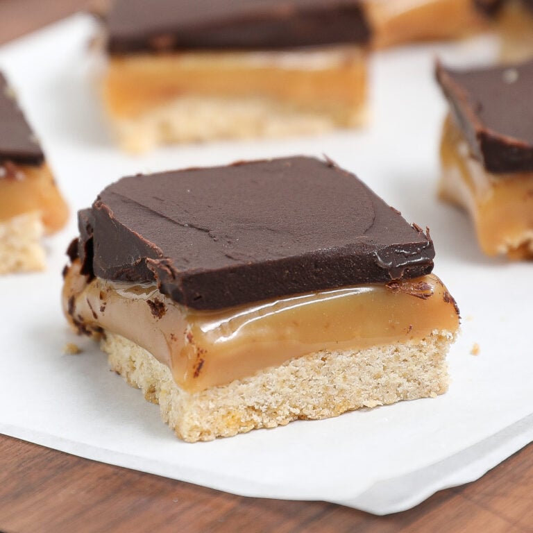 A close-up of millionaire bars, featuring a caramel shortbread bar topped with a thick layer of chocolate, gooey caramel in the middle, and a crumbly cookie base, resting on white parchment paper.