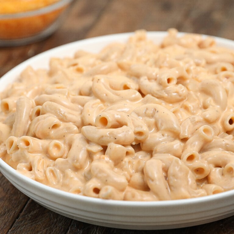 A white bowl filled with creamy Stove Top Mac and Cheese sits on a wooden table, with a blurred dish of shredded cheese in the background.
