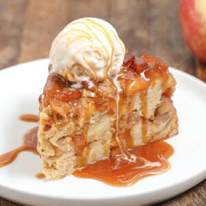 A slice of caramel apple bread pudding topped with vanilla ice cream and drizzled with caramel sauce, served on a white plate with an apple in the background.