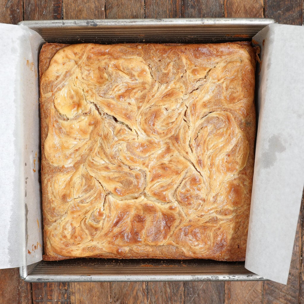 A square pan lined with parchment paper holds golden-brown carrot cake brownie bars with a beautifully swirled top, sitting on a rustic wooden surface.