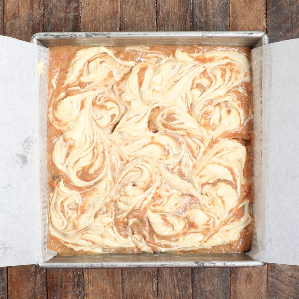 A square baking pan lined with parchment paper holds unbaked carrot cake brownie bars, their swirled pattern of light and caramel-colored batter on top, resting on a rustic wooden surface.