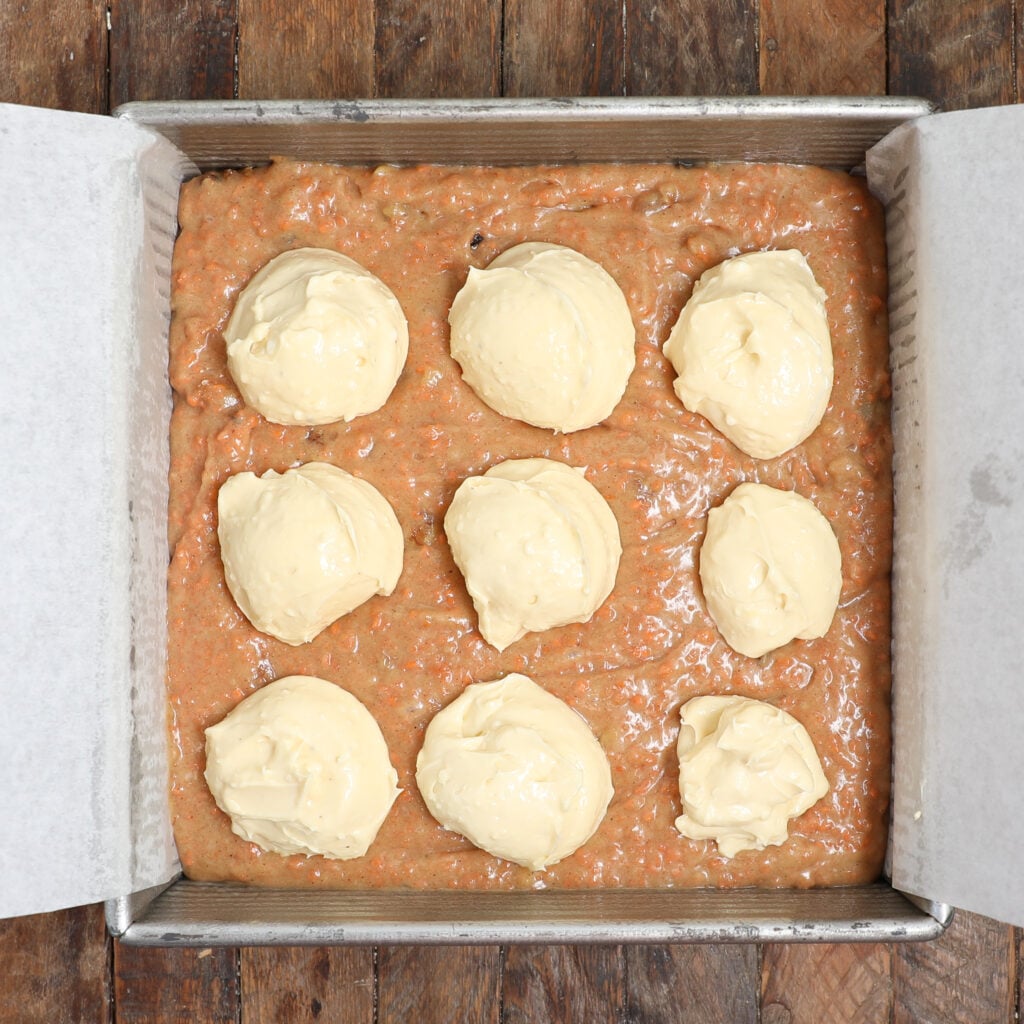 A square metal baking pan lined with parchment paper holds unbaked carrot cake brownie bars, with nine dollops of cream cheese mixture evenly spaced on top. The pan sits on a rustic wooden surface.