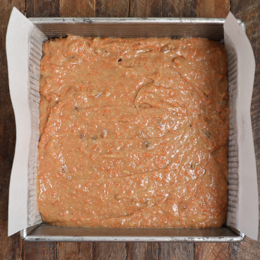 A rectangular metal baking pan lined with parchment paper, filled with raw carrot cake brownie bars batter speckled with orange shreds, sits on a wooden surface.