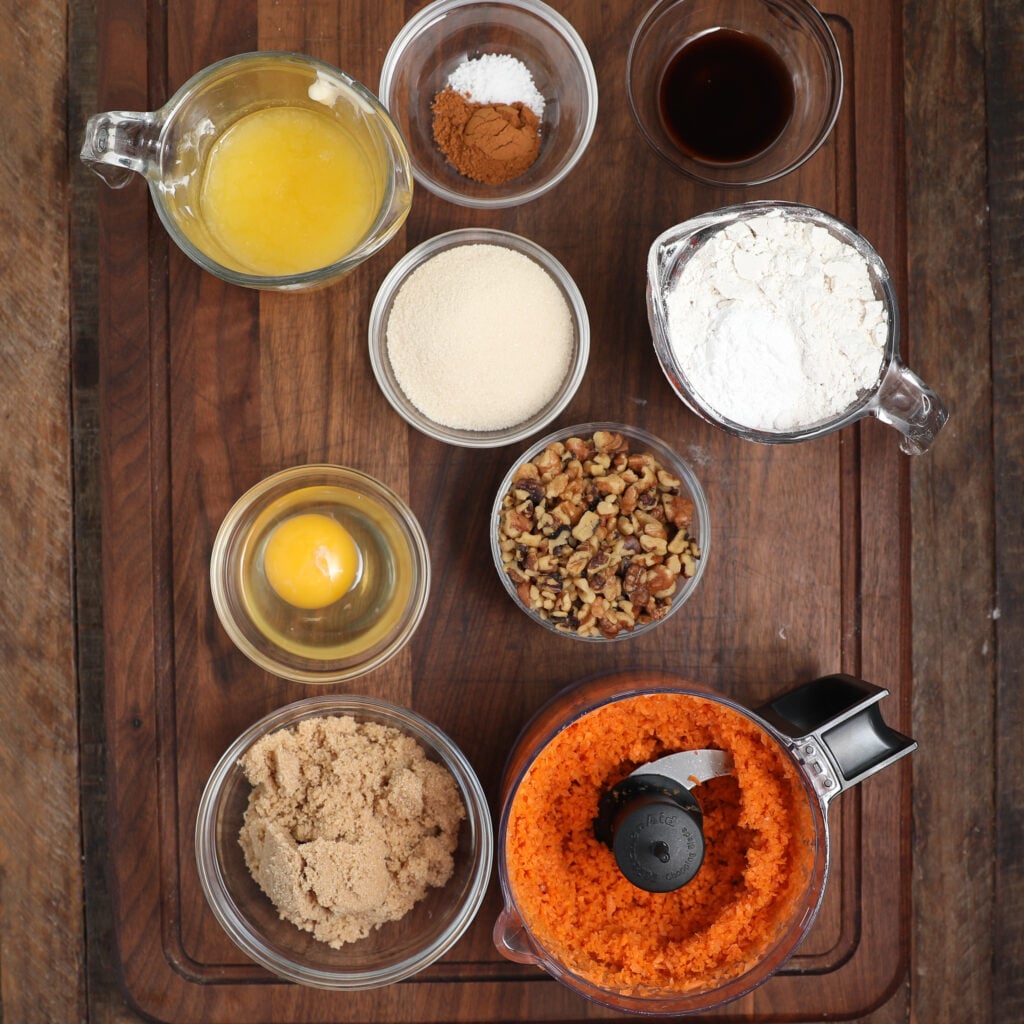 Top-down view of baking ingredients for carrot cake brownie bars on a wooden board: melted butter, spices, vanilla, flour, breadcrumbs, egg, chopped walnuts, brown sugar, and grated carrots in a food processor.