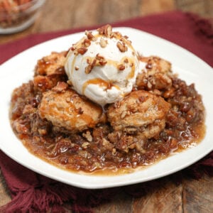 A dessert plate with warm pecan pie cobbler topped with vanilla ice cream, drizzled with caramel sauce, and sprinkled with chopped pecans, served on a rustic wooden table with a red cloth napkin.