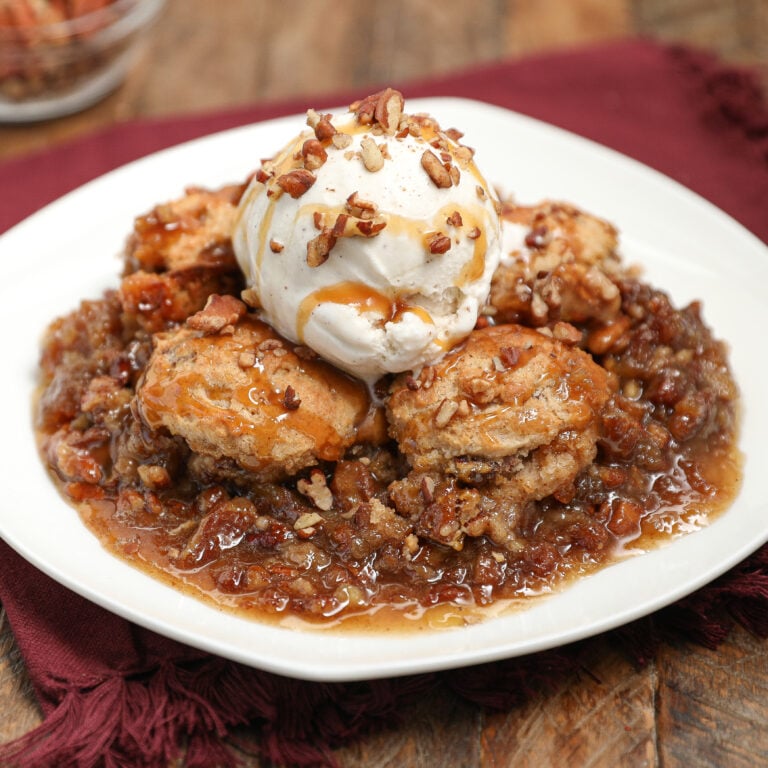 A dessert plate with warm pecan pie cobbler topped with vanilla ice cream, drizzled with caramel sauce, and sprinkled with chopped pecans, served on a rustic wooden table with a red cloth napkin.