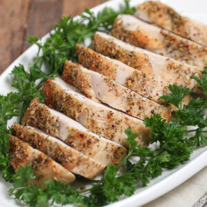 Sliced, herb-seasoned roasted turkey breast and chicken breast are arranged neatly on a white plate, garnished with fresh parsley.
