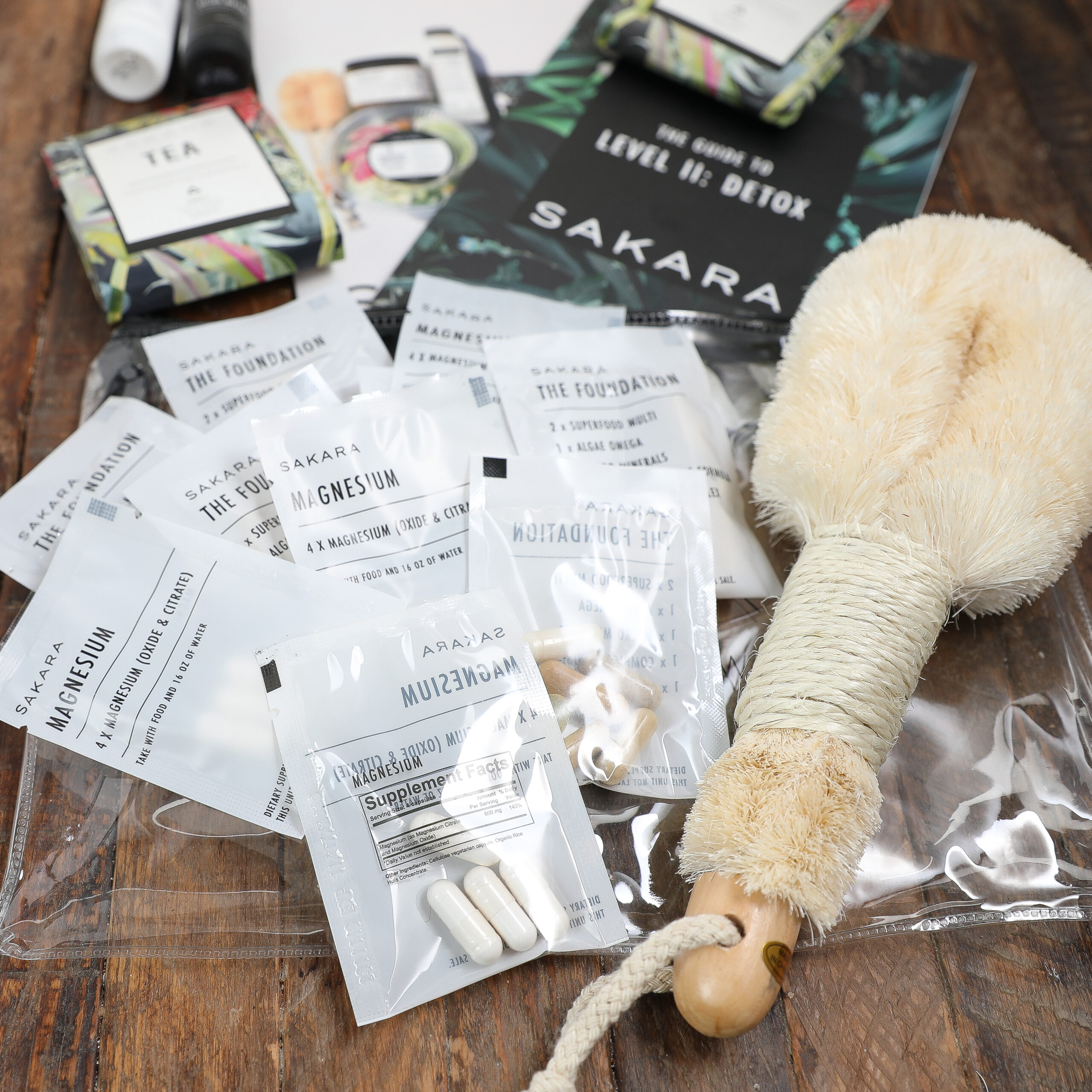 A collection of Sakara Level wellness products—including supplement packets, tea sachets, and a dry body brush—arranged on a wooden surface. Some packets are labeled magnesium and multivitamin.